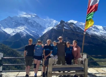 Group of trekkers posing by railing with prayer flag and snowy Annapurna peaks behind.
